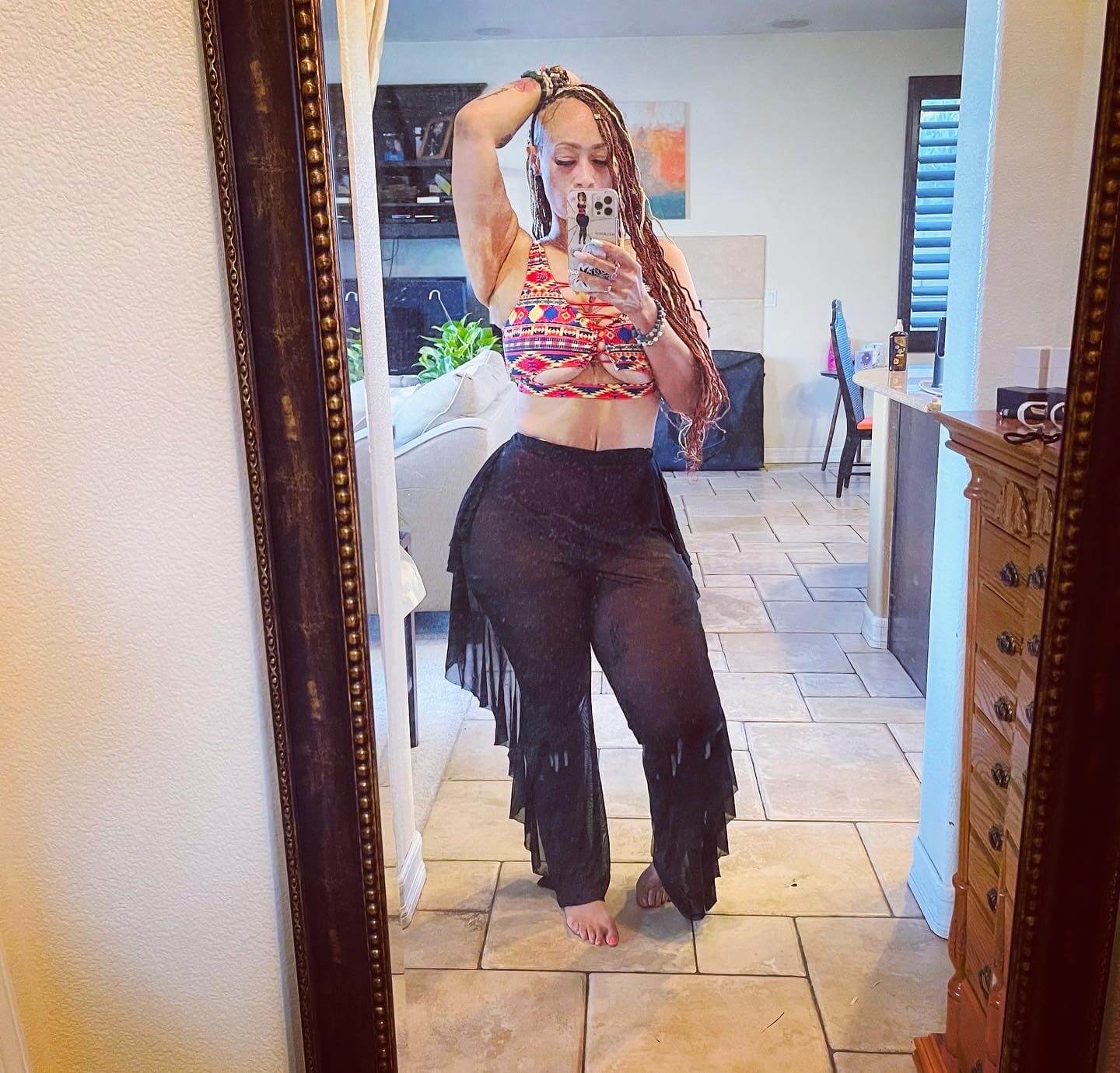 Woman taking a mirror selfie at home, showing off toned figure in colorful crop top and sheer black pants