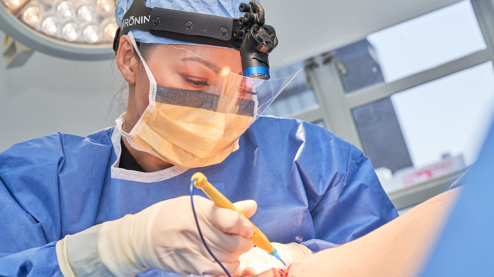 Plastic surgeon performing surgery wearing protective surgical gear in a brightly lit operating room.