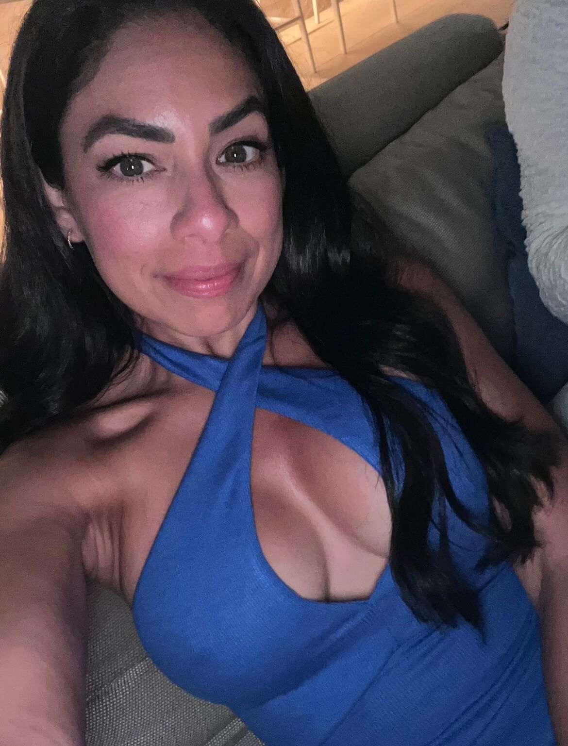 Woman smiling gently, with long dark hair and wearing a stylish blue cross-neck top.