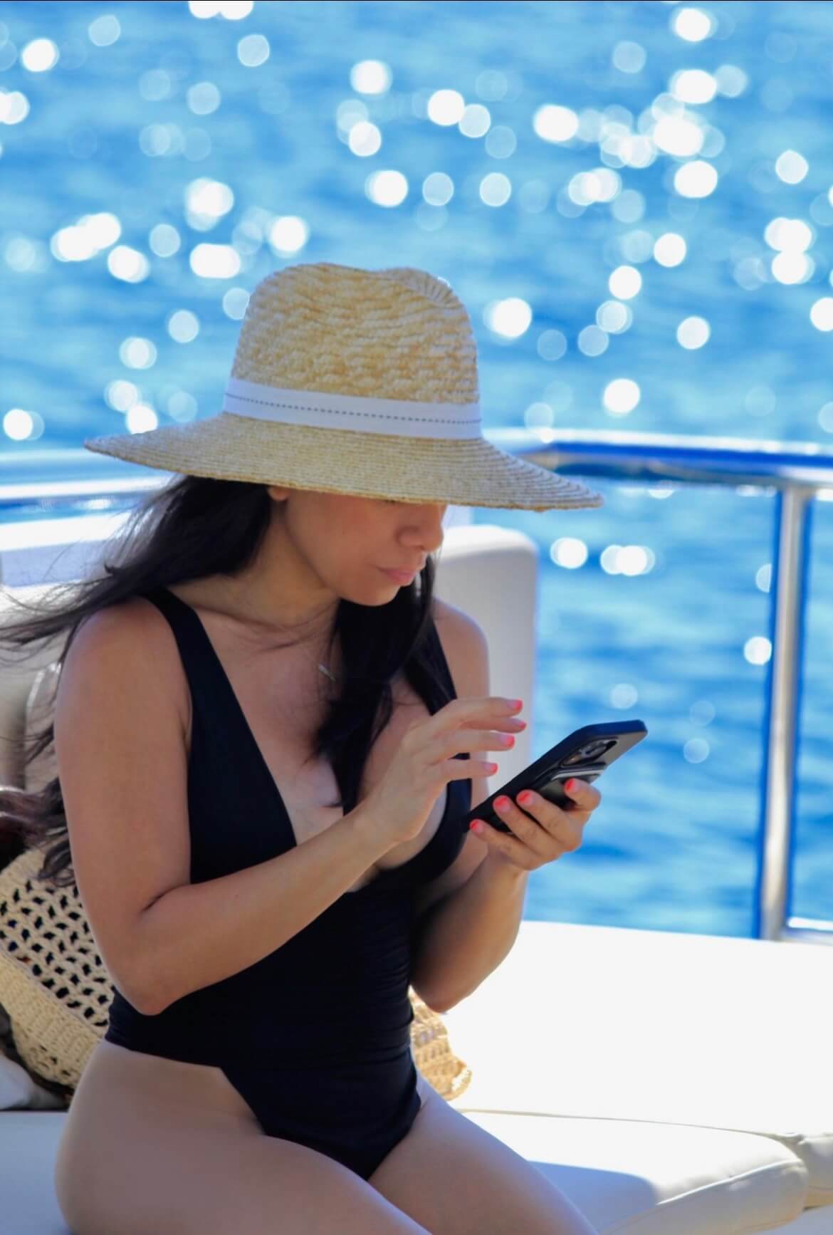 Woman wearing a black swimsuit and straw hat relaxing on a boat while using a smartphone.