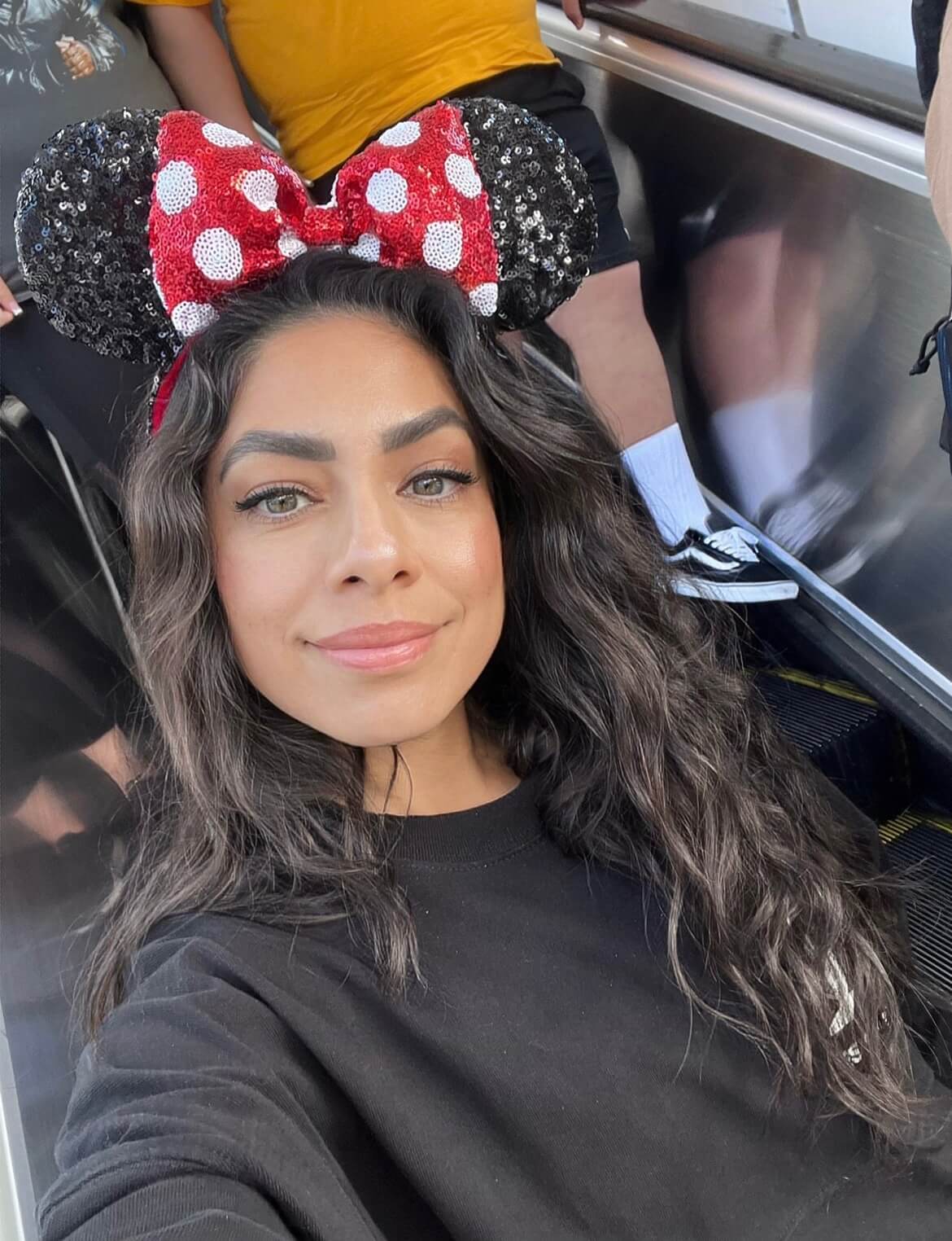 Smiling woman with long dark hair wearing a sequined Minnie Mouse ears headband and black sweater.