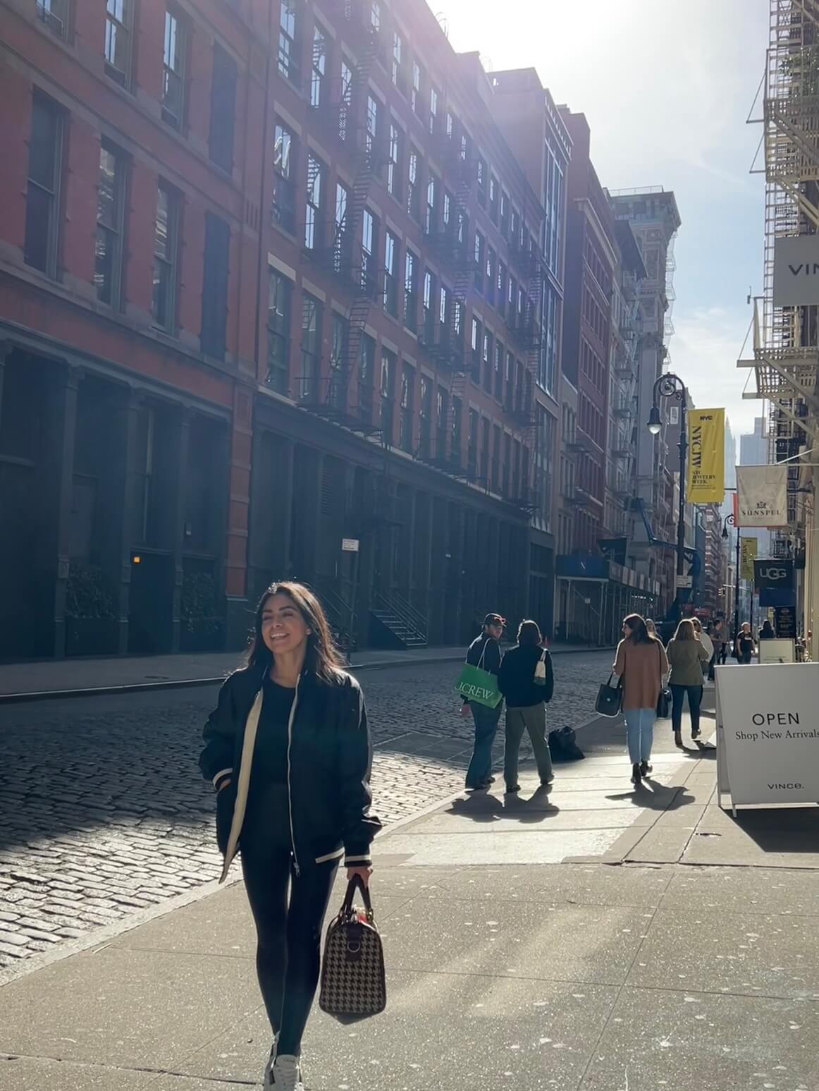 Woman walking down a sunlit street in SoHo, New York, smiling and carrying a designer bag