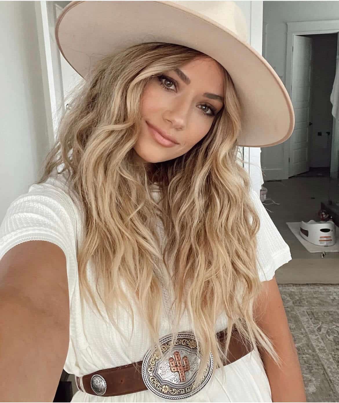 Woman with long blonde wavy hair wearing a wide-brimmed hat and white dress with a decorative belt, taking a selfie indoors.
