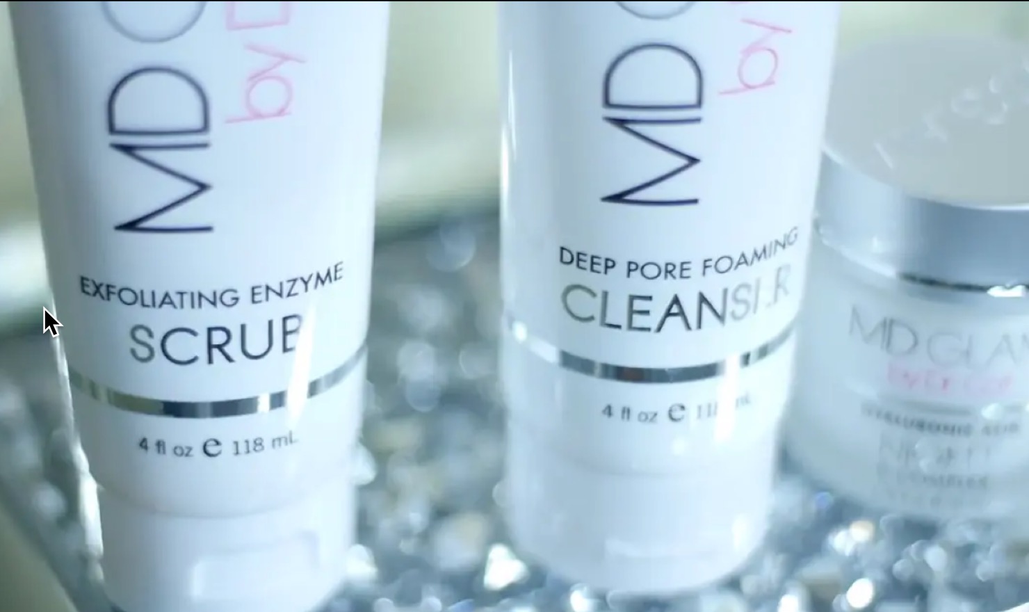 Close-up image of MD Glam skincare products including Exfoliating Enzyme Scrub and Deep Pore Foaming Cleanser.