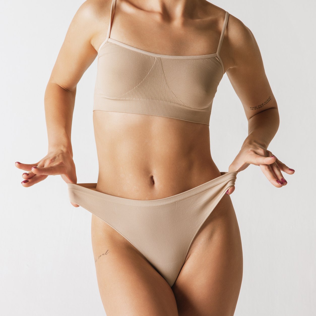 Woman in a beige bra adjusting the waistband of her briefs, showing a trim waist and hips