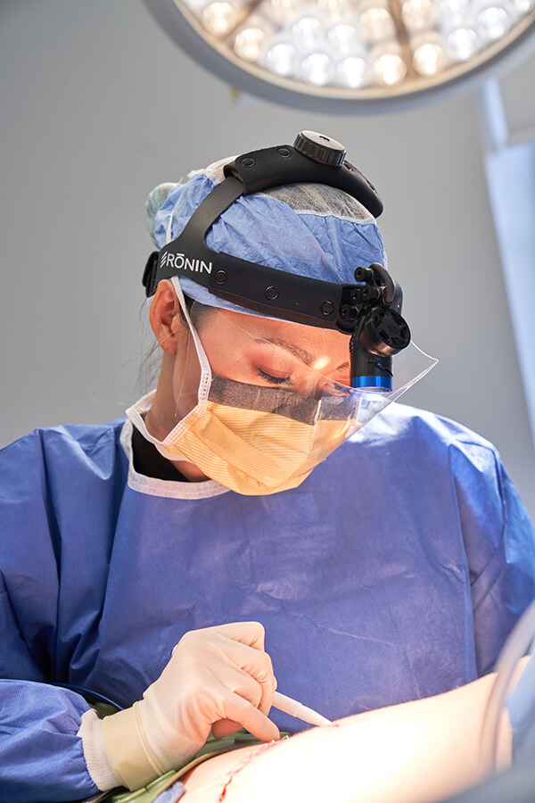 Dr. Cat Begovic in surgical loupes and gown closing an incision in the operating room